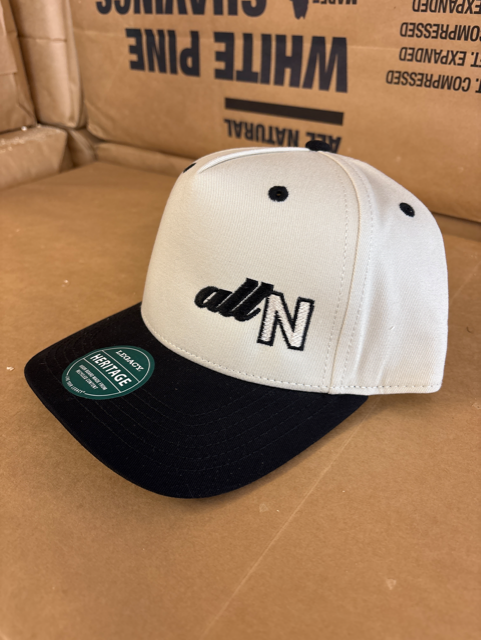 A structured baseball cap with a white crown and black brim, featuring the text 'alt N' embroidered on the front. A green circular sticker is visible on the brim, and the cap is resting on a brown cardboard box.