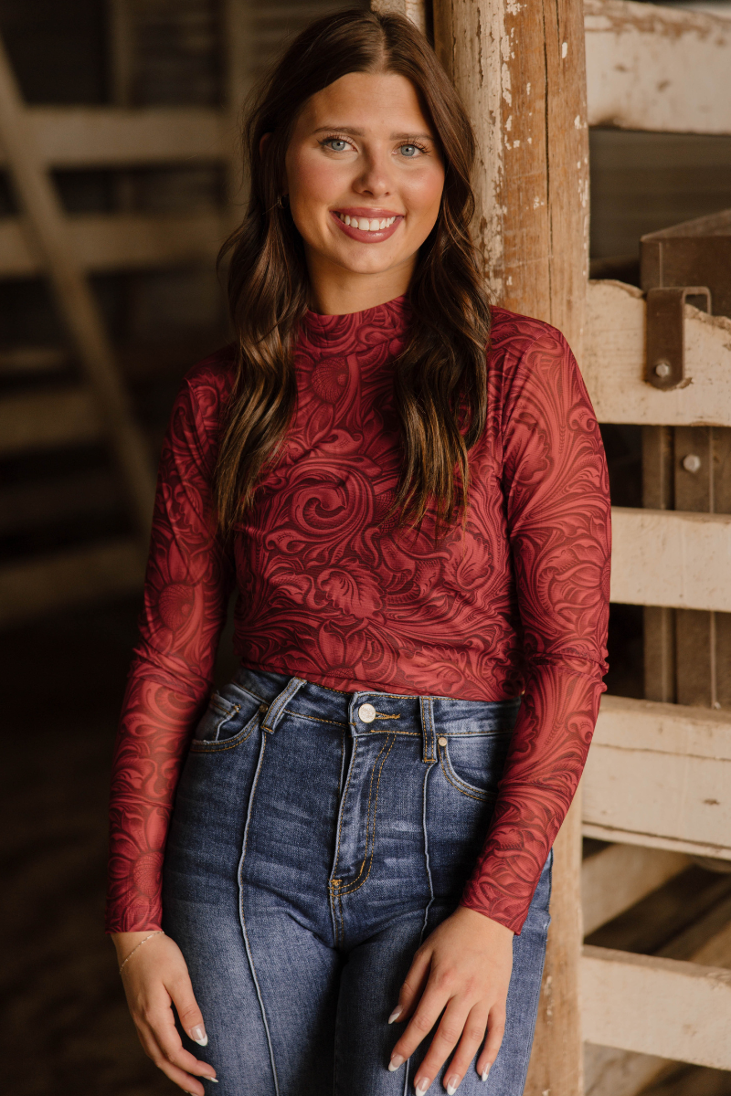 Woman wearing a red patterned long-sleeve top and blue jeans standing in a rustic setting.