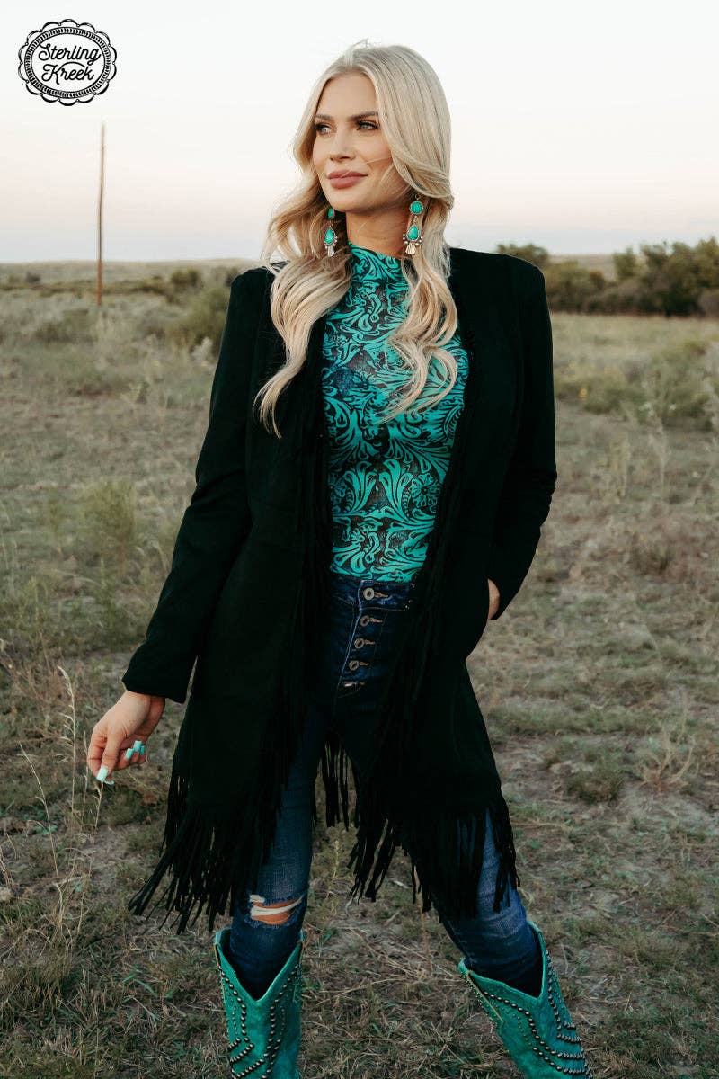 Woman in a black coat with fringe, teal patterned top, and blue jeans standing in a field.
