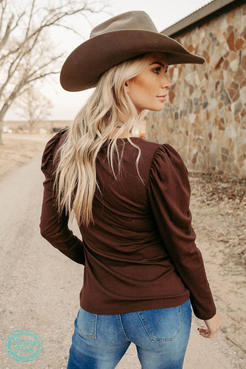 Woman wearing a brown cowboy hat and long-sleeve top, standing outdoors.