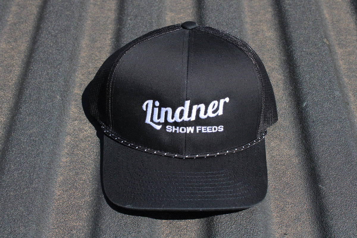 A black trucker-style cap with white embroidered text reading 'Lindner SHOW FEEDS' on the front, photographed against a corrugated gray metal background.