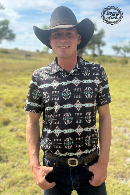 Man wearing a black patterned shirt and cowboy hat in an outdoor setting