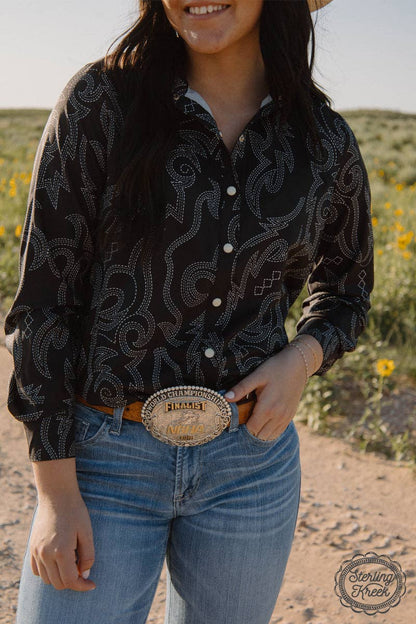 Person wearing a black patterned shirt and blue jeans with a large belt buckle in a desert setting.