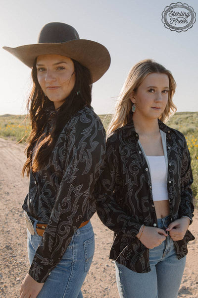 Two women in patterned shirts and jeans standing on a dirt road with a clear sky.