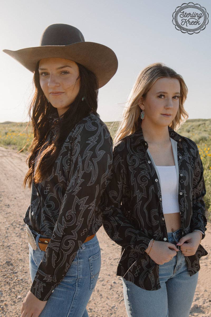 Two women in patterned shirts and jeans standing on a dirt road with a clear sky.