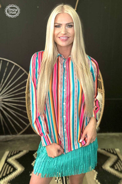 Woman wearing a colorful striped shirt and turquoise fringe skirt in front of a decorative wall.