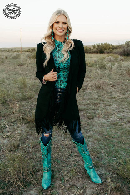 Woman in a black fringe jacket and turquoise boots standing in a field.
