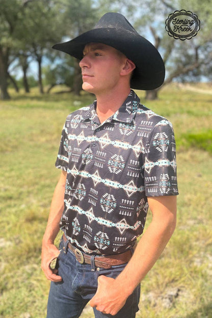 Man wearing a black cowboy hat and patterned shirt standing in a grassy field.
