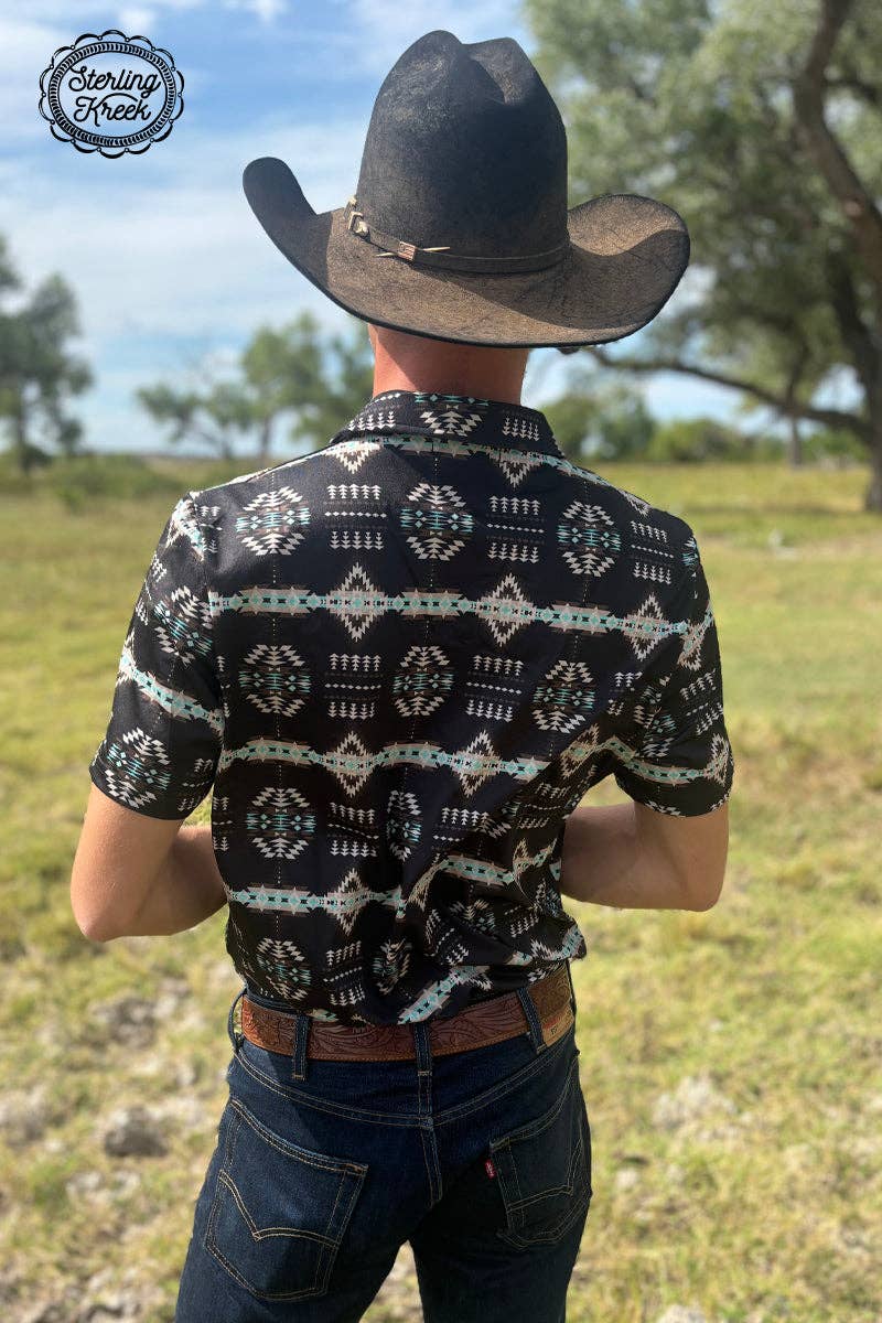 Man wearing a black cowboy hat and patterned shirt standing in a grassy field.