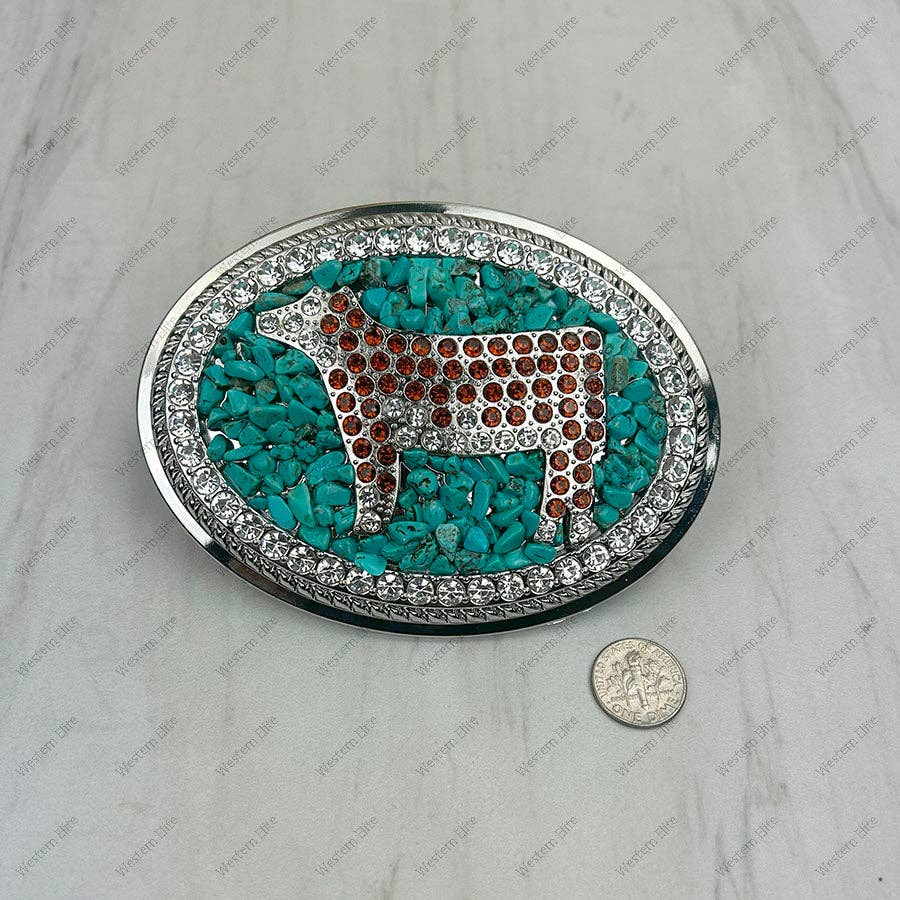 An oval silver-tone belt buckle featuring a cow design paved with brown and clear rhinestones, surrounded by a border of turquoise and clear crystals, shown on a light marble surface with a coin for scale.
