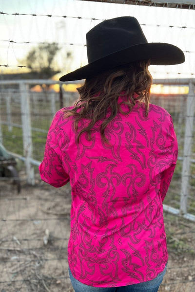 Person wearing a bright pink long-sleeve shirt with black patterns and a black cowboy hat, standing in front of a barbed wire fence.
