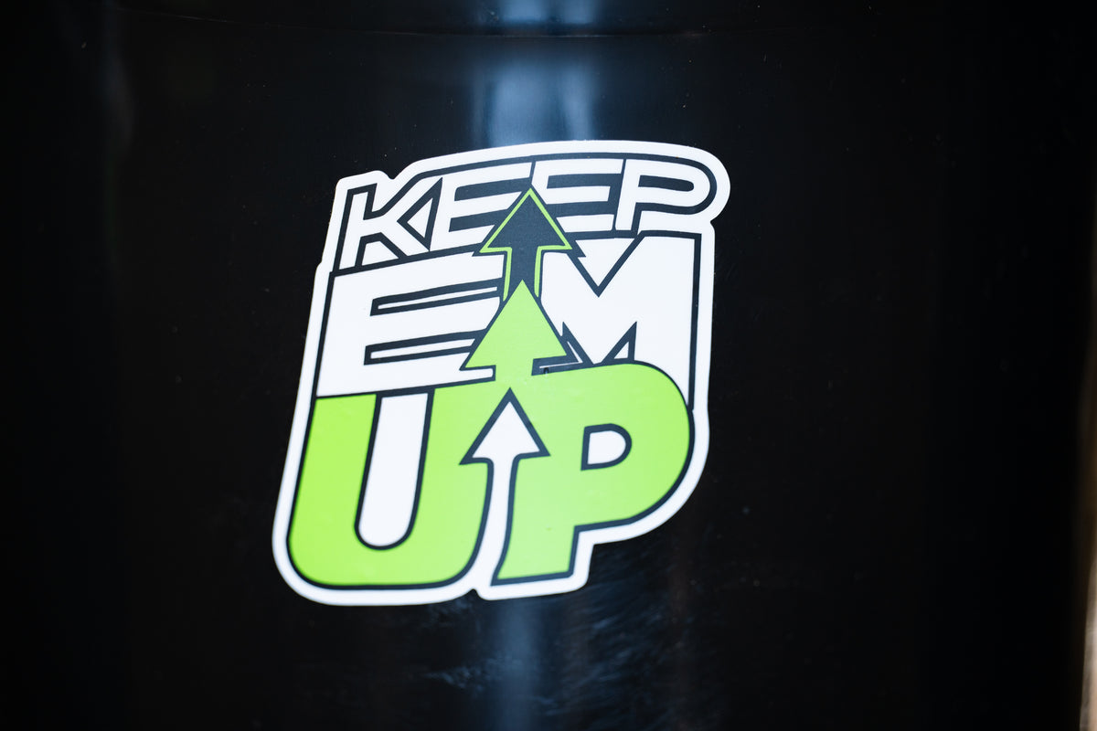 A close-up of a die-cut sticker on a black surface. The sticker features the words 'KEEP EM UP' in stylized lettering, with a bright green upward-pointing arrow layered through the design.