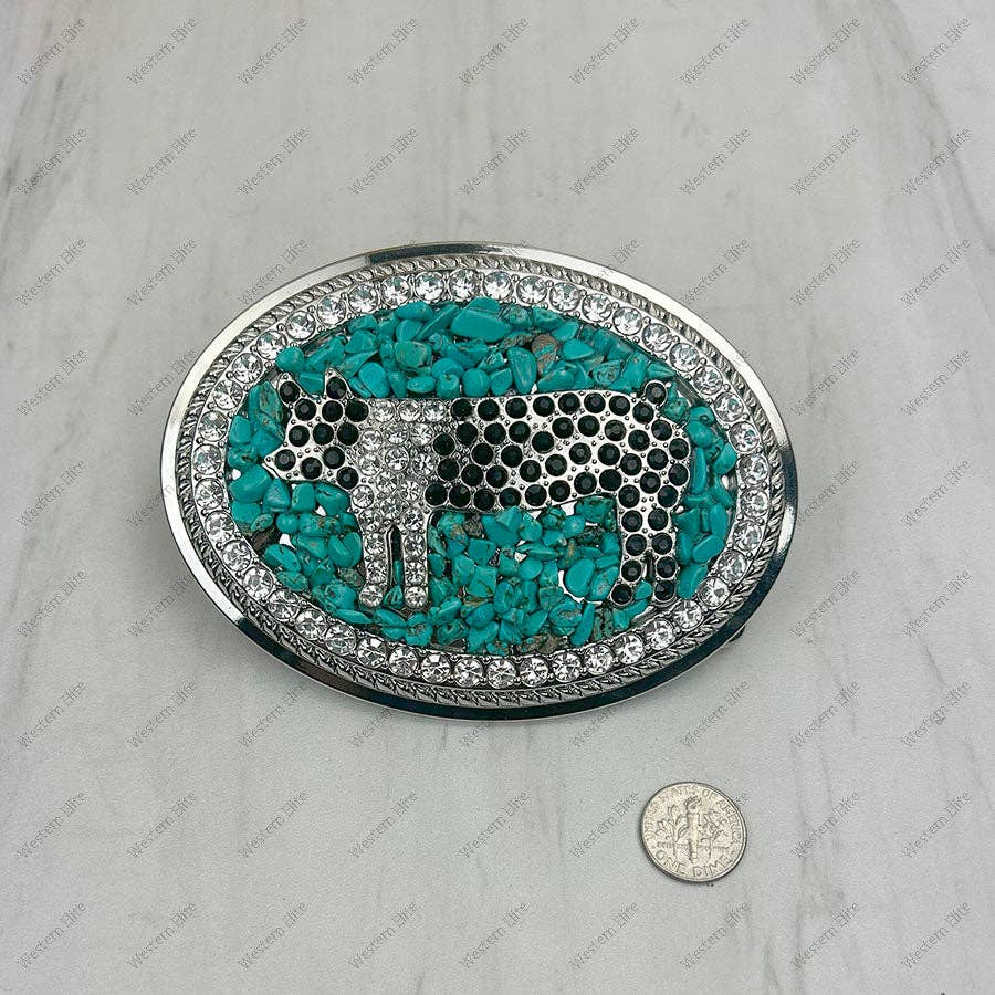 An oval silver-toned belt buckle featuring a central turquoise and black-and-white spotted animal motif, surrounded by a border of clear rhinestones, shown on a light marble-like surface with a coin for scale.