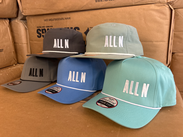 Four baseball caps in different colors (gray, black, blue, and mint green) arranged on a cardboard surface with brown shipping boxes in the background. Each cap features the text 'ALL N' embroidered on the front in white.