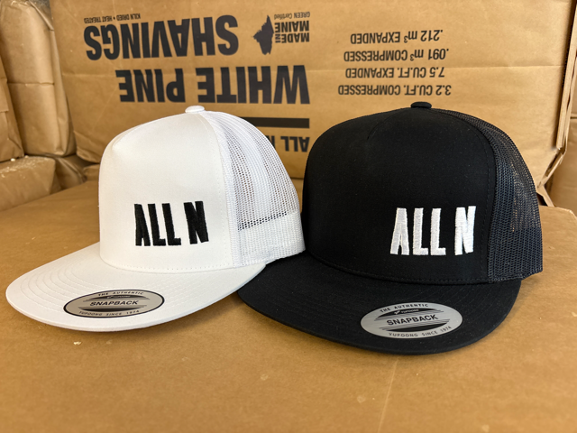 Two snapback trucker hats displayed side by side on a cardboard surface, with a large cardboard box in the background. The left hat is white with black mesh back and black 'ALL N' text on the front.