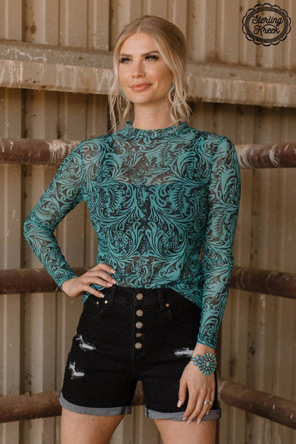 Woman wearing a teal patterned top and black shorts standing against a wooden background