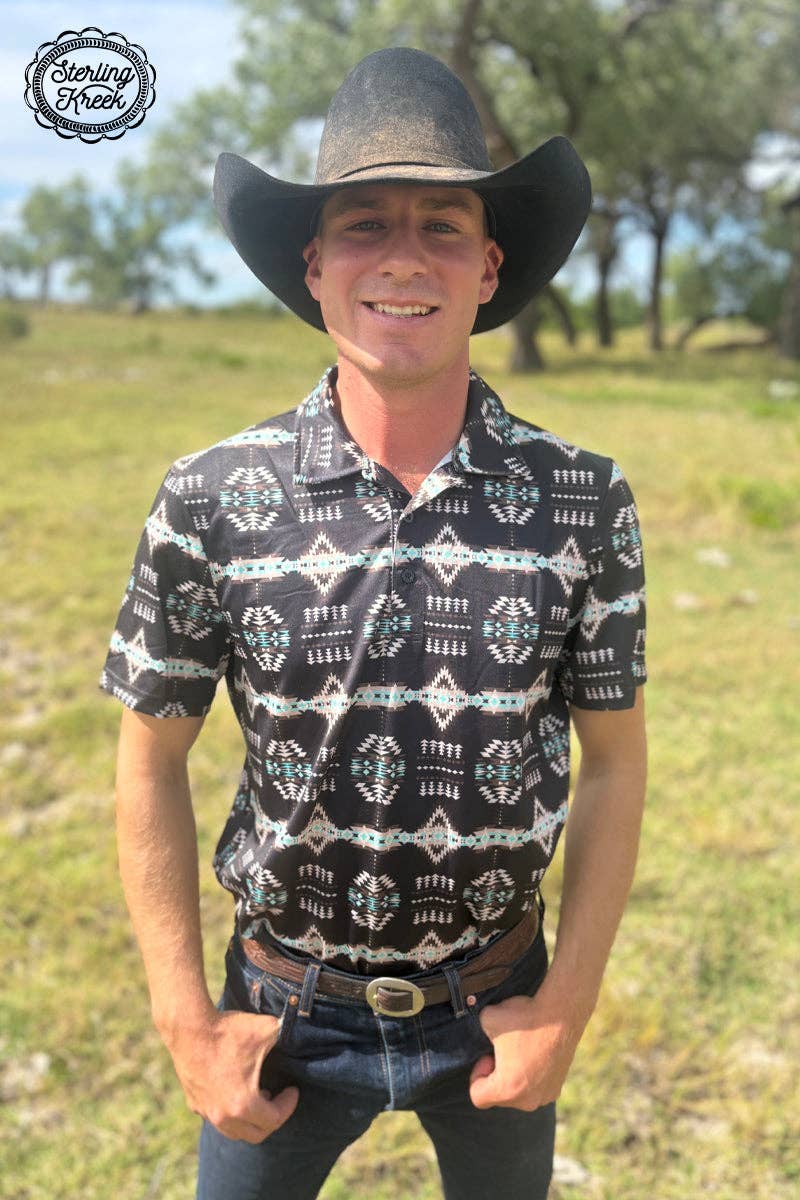 Man wearing a black cowboy hat and patterned shirt standing in a grassy field.