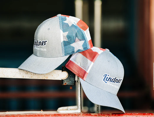 Two baseball caps with American flag design and 'Lindner' branding on a blurred background.