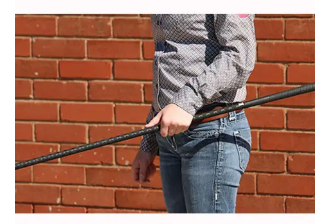 A person wearing a gray patterned long-sleeve shirt and blue jeans holds a long black pole or rod in front of a red brick wall.